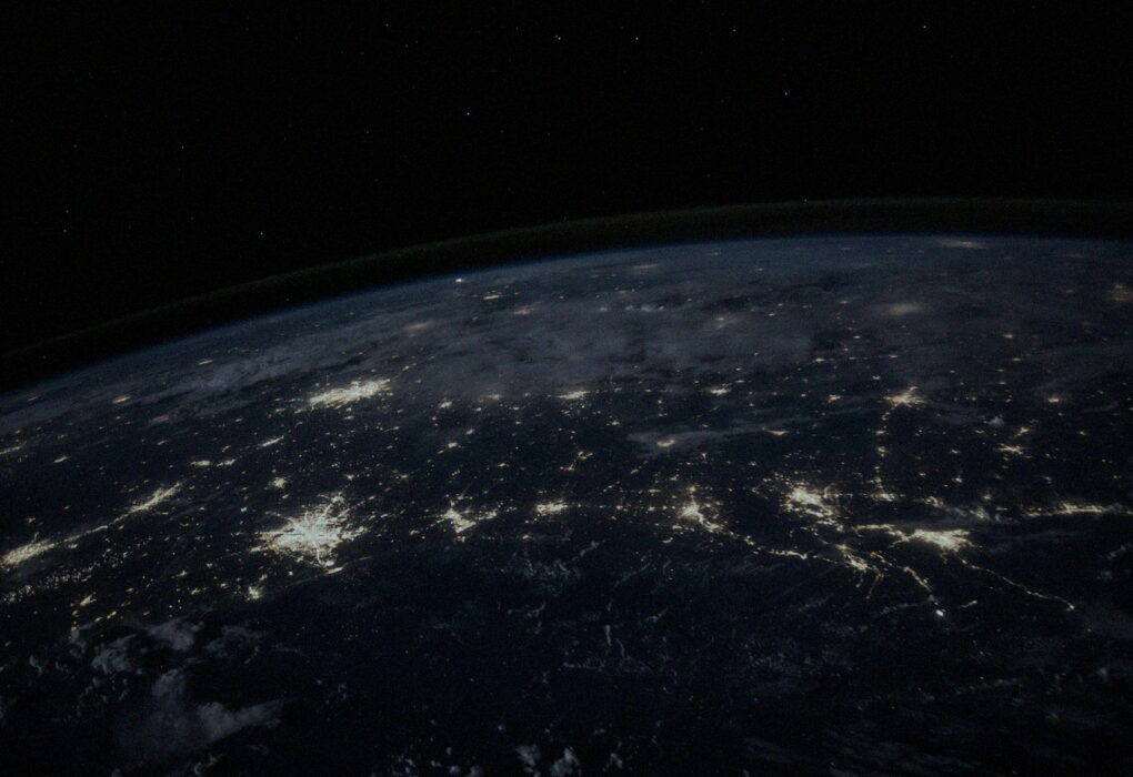 Earth at night from orbit with glowing city-light networks along the curved horizon, no people or brand logos — global interconnected finance and cyber mood