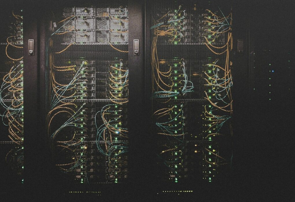Dark data-center server racks behind mesh doors with teal and orange cabling and green status LEDs, no people, used as a still for long-running AI agent infrastructure