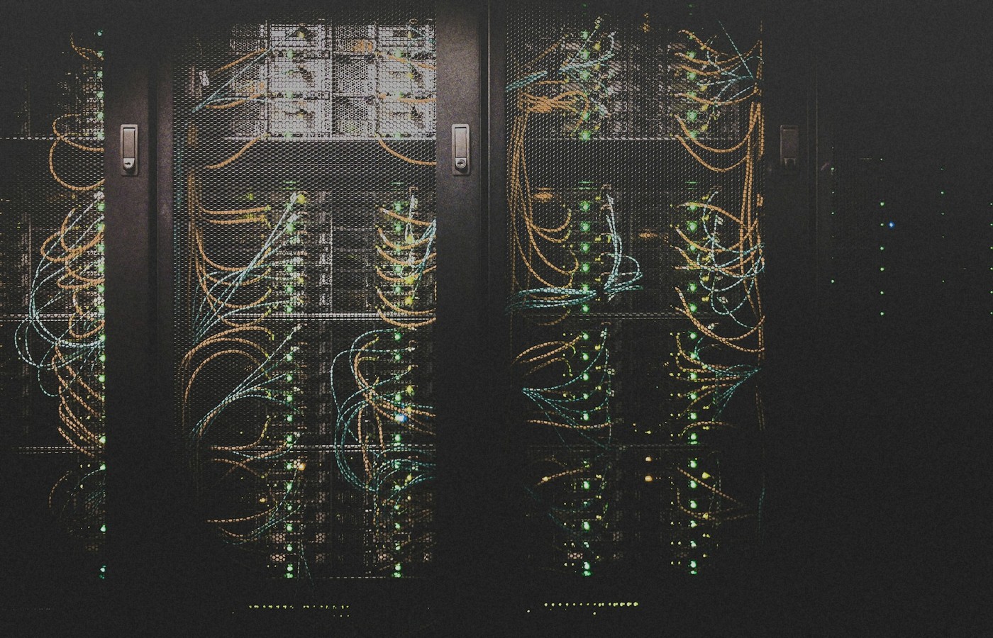 Dark data-center server racks behind mesh doors with teal and orange cabling and green status LEDs, no people, used as a still for long-running AI agent infrastructure