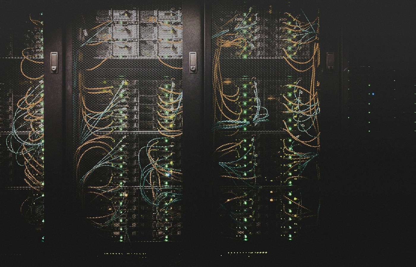 Dark datacenter server racks behind mesh doors with dense yellow cable loops and green status LEDs, face-free infrastructure still for Gimlet multi-silicon coverage