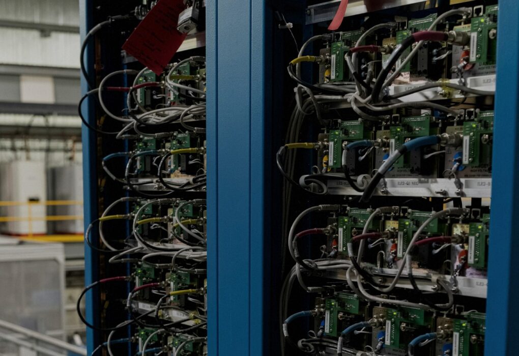 Close-up of two blue industrial server racks packed with green circuit modules and dense multi-color cabling in a face-free facility still for iPronics OCS coverage
