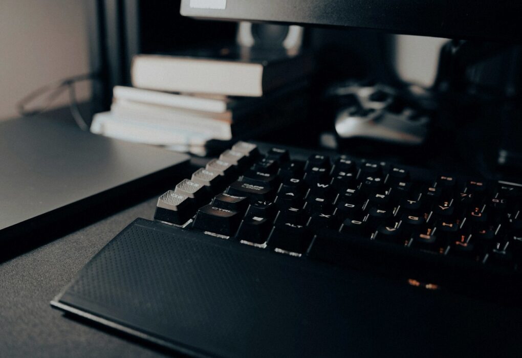 Face-free close-up of a black mechanical keyboard on a dark desk with orange key lighting and notebooks in soft focus — coding desk still for Cognition Series E Devin coverage