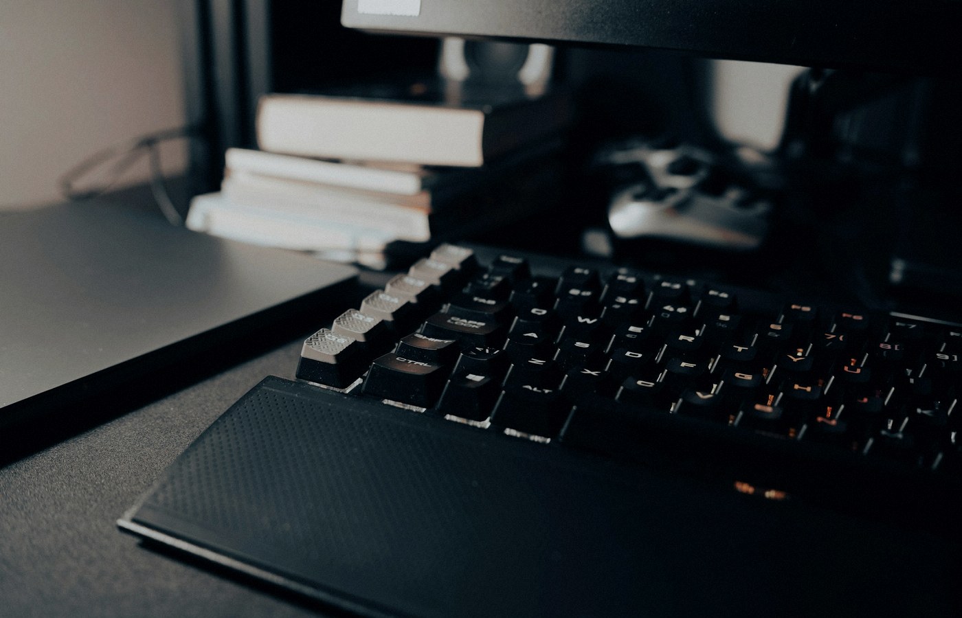 Face-free close-up of a black mechanical keyboard on a dark desk with orange key lighting and notebooks in soft focus — coding desk still for Cognition Series E Devin coverage