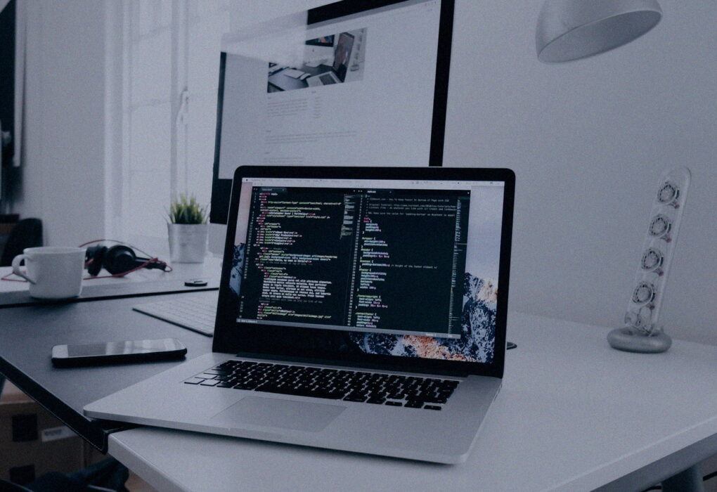 Face-free MacBook with code on screen beside a phone and headphones on a white desk — laptop workspace still for Meta Muse personal AI agent launch coverage