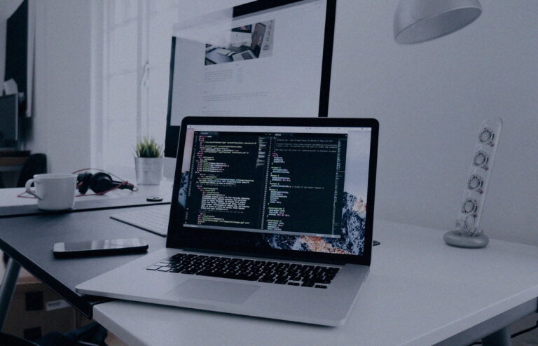 Face-free MacBook with code on screen beside a phone and headphones on a white desk — laptop workspace still for Meta Muse personal AI agent launch coverage