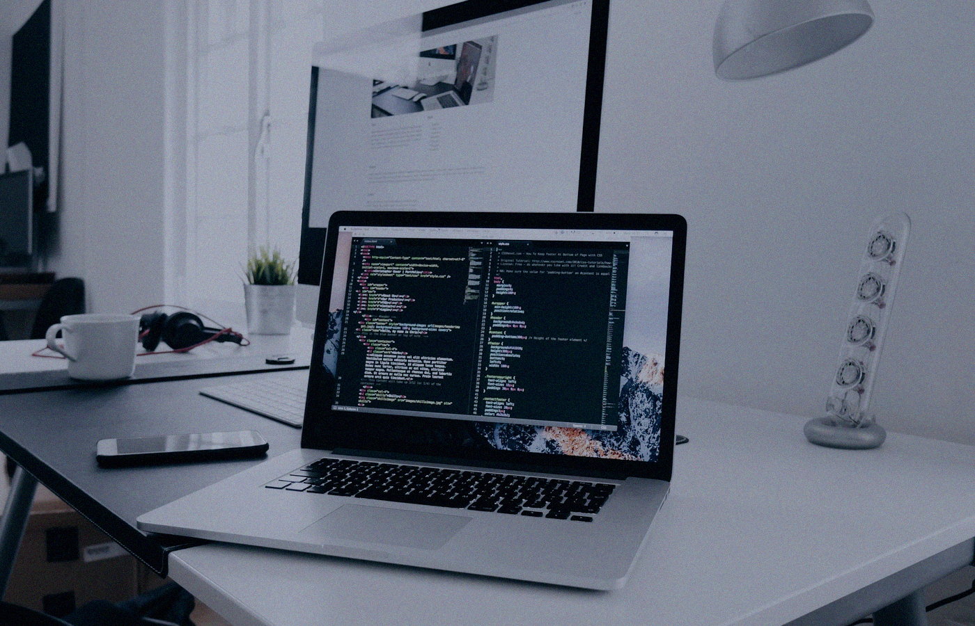 Face-free MacBook with code on screen beside a phone and headphones on a white desk — laptop workspace still for Meta Muse personal AI agent launch coverage