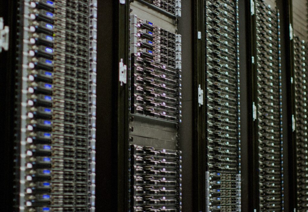 Close-up of face-free data-center server racks with rows of blue status LEDs receding into soft focus — AI cloud compute still for Nscale × Figure coverage