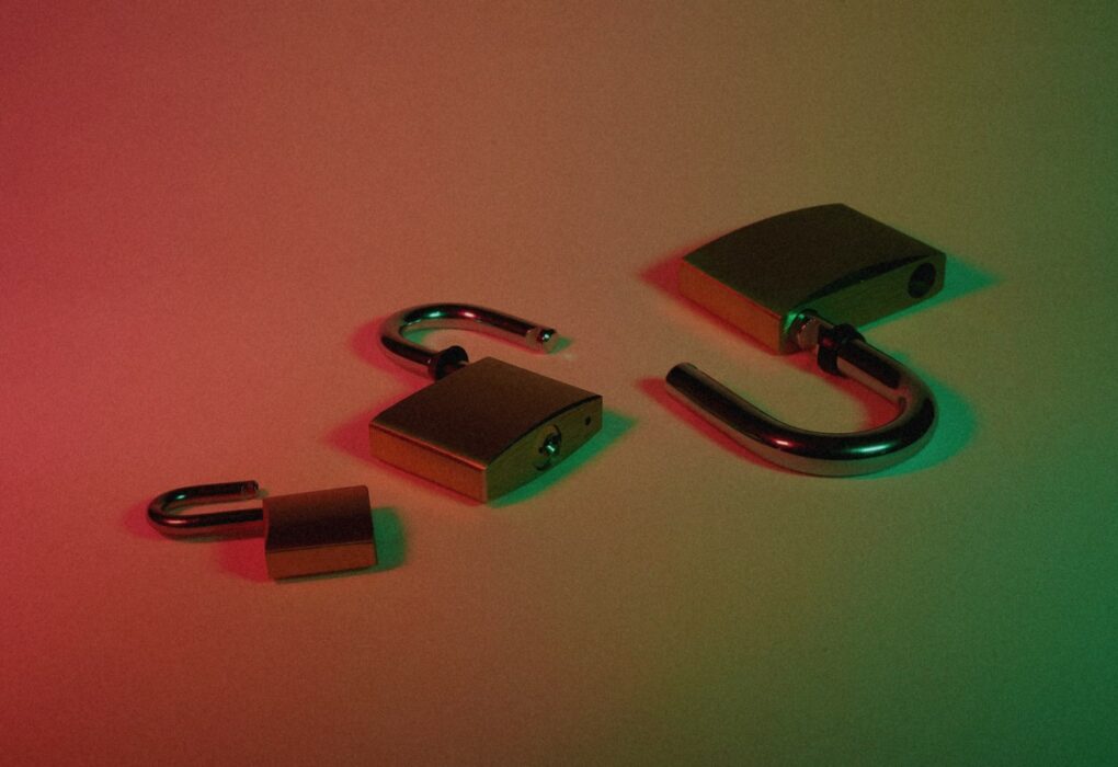Three open brass padlocks on a surface under red-to-green dramatic lighting, face-free still for OpenAI Astra Critical cybersecurity capability
