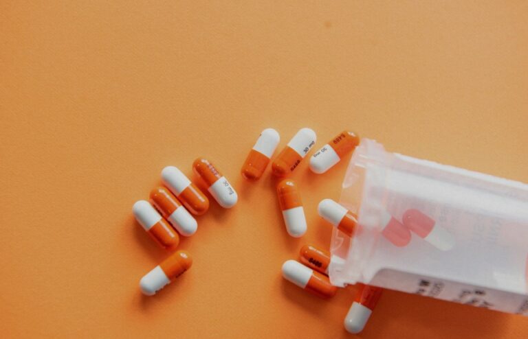 Face-free orange-and-white medication capsules spilled from a translucent prescription bottle on an orange surface — pharmacy and Rx still for Forus Series C AI medication-access coverage