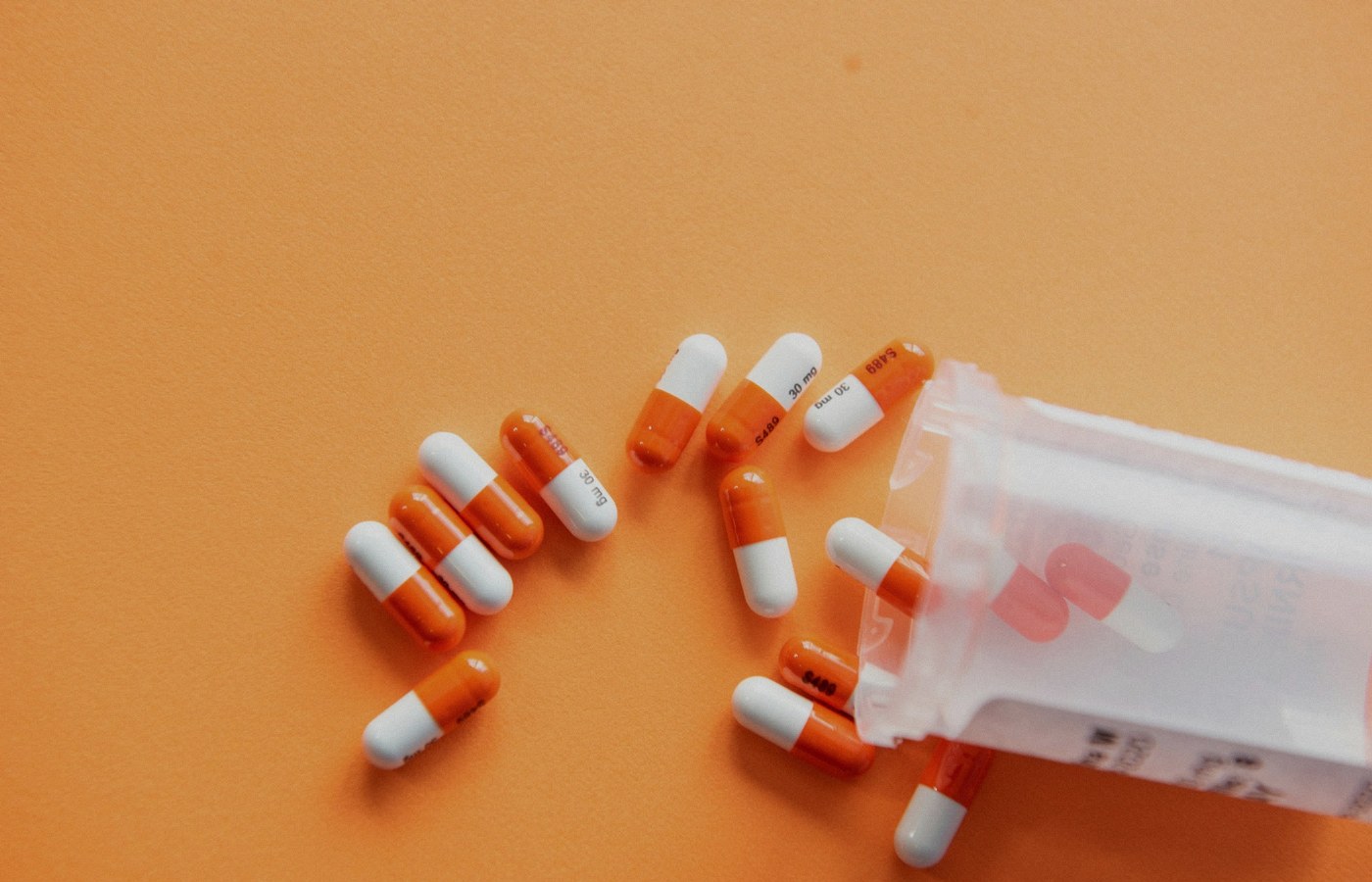 Face-free orange-and-white medication capsules spilled from a translucent prescription bottle on an orange surface — pharmacy and Rx still for Forus Series C AI medication-access coverage