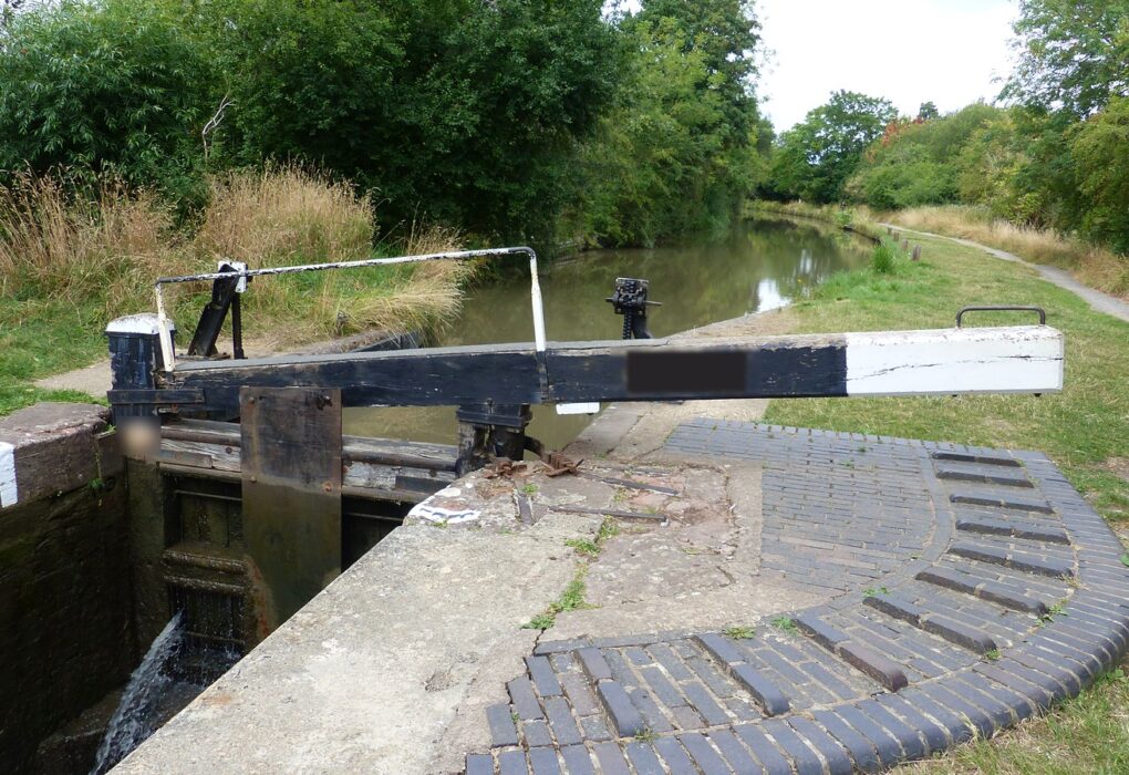 A black-and-white canal lock beam over a closed lock gate, water spilling below, brick footing and a grassy towpath