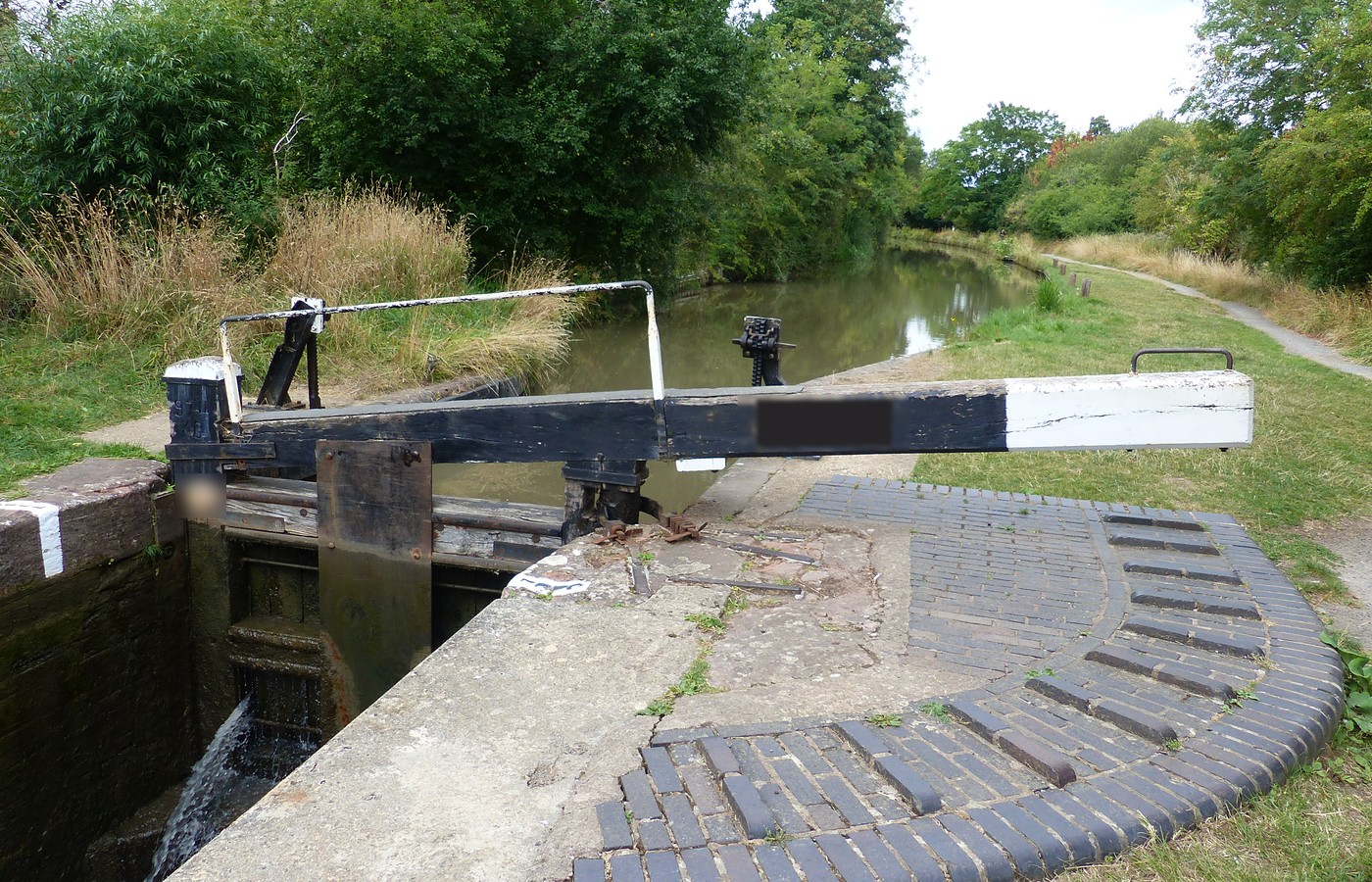 A black-and-white canal lock beam over a closed lock gate, water spilling below, brick footing and a grassy towpath