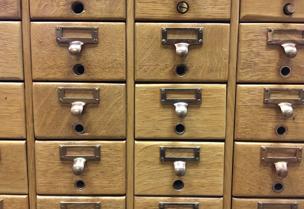 A wall of oak card-catalog drawers in neat rows, each with an empty brass label holder, a curved pull and a small rod hole