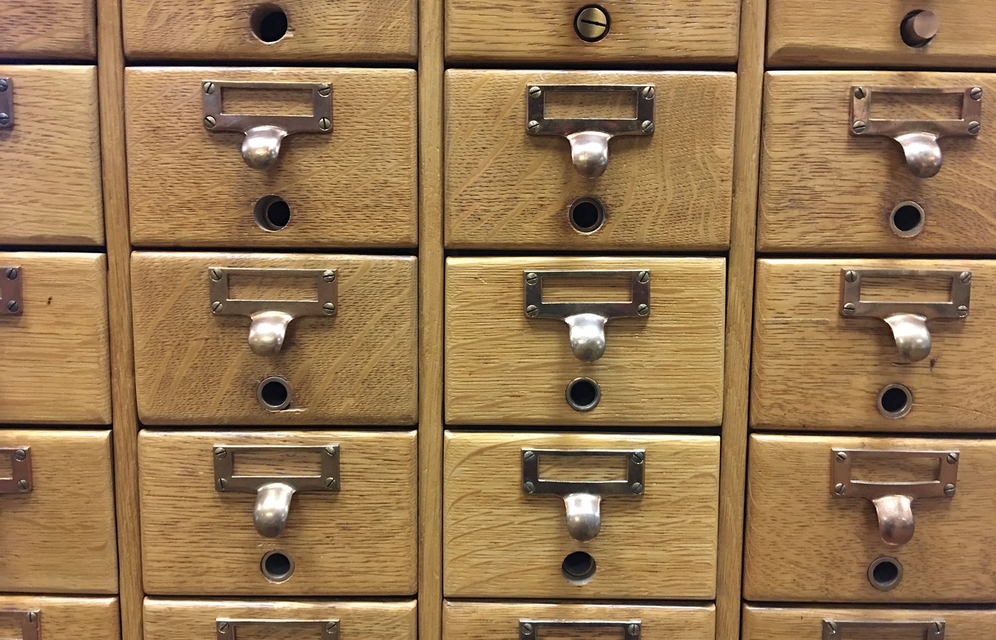 A wall of oak card-catalog drawers in neat rows, each with an empty brass label holder, a curved pull and a small rod hole