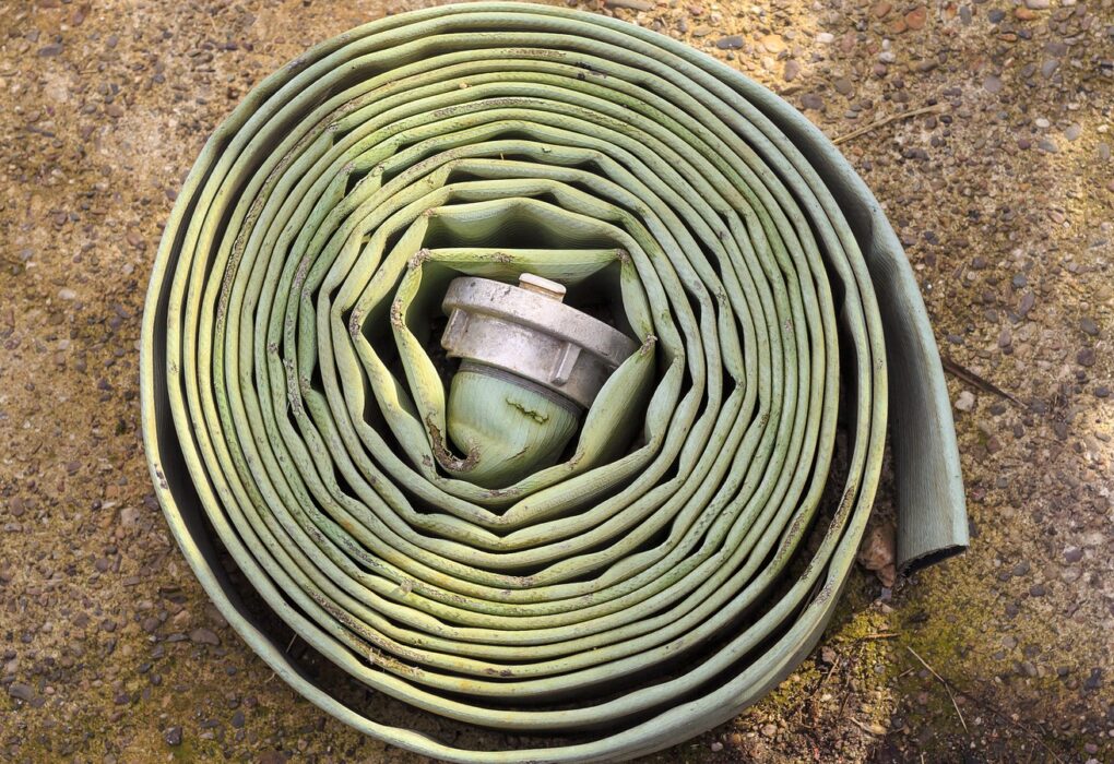 A worn, pale green flat fire hose wound into a tight coil on gravelly ground, with a plain metal coupling tucked in the center and no people in frame