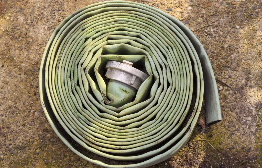 A worn, pale green flat fire hose wound into a tight coil on gravelly ground, with a plain metal coupling tucked in the center and no people in frame