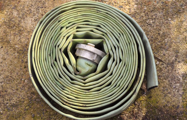 A worn, pale green flat fire hose wound into a tight coil on gravelly ground, with a plain metal coupling tucked in the center and no people in frame