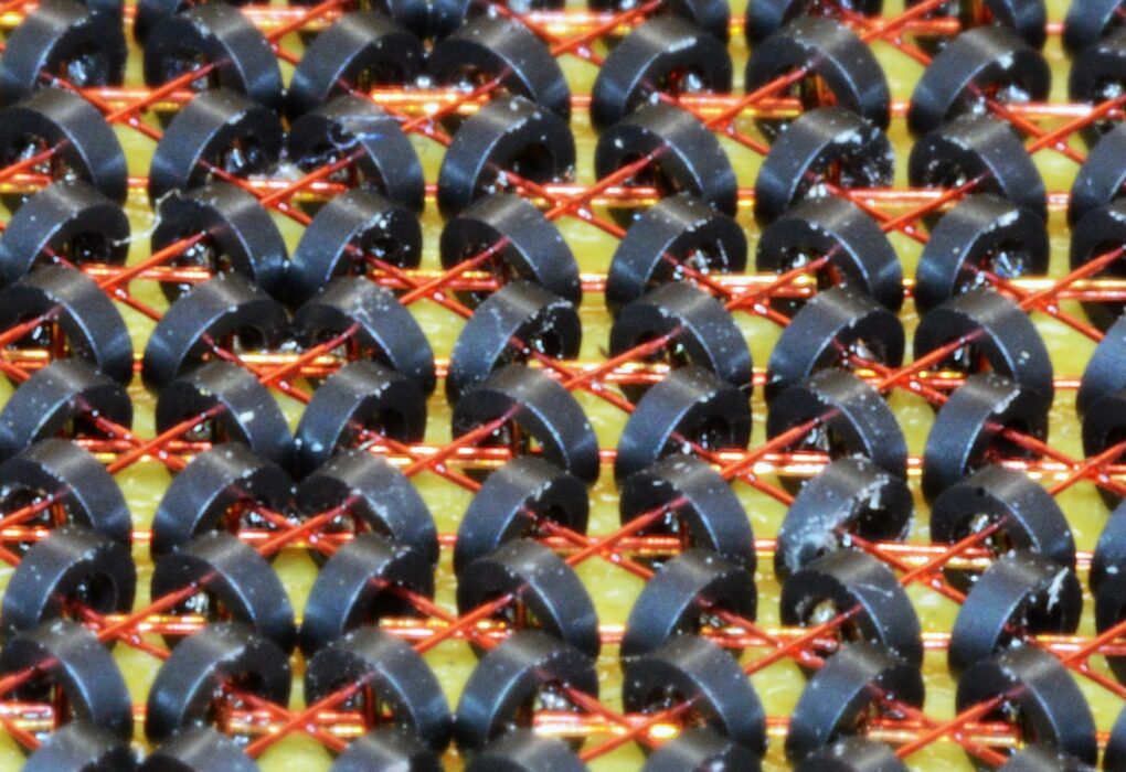 Close-up of a magnetic-core memory plane: rows of small dark ferrite rings threaded on thin copper wires over a yellow board