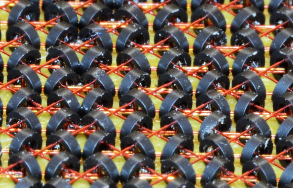 Close-up of a magnetic-core memory plane: rows of small dark ferrite rings threaded on thin copper wires over a yellow board