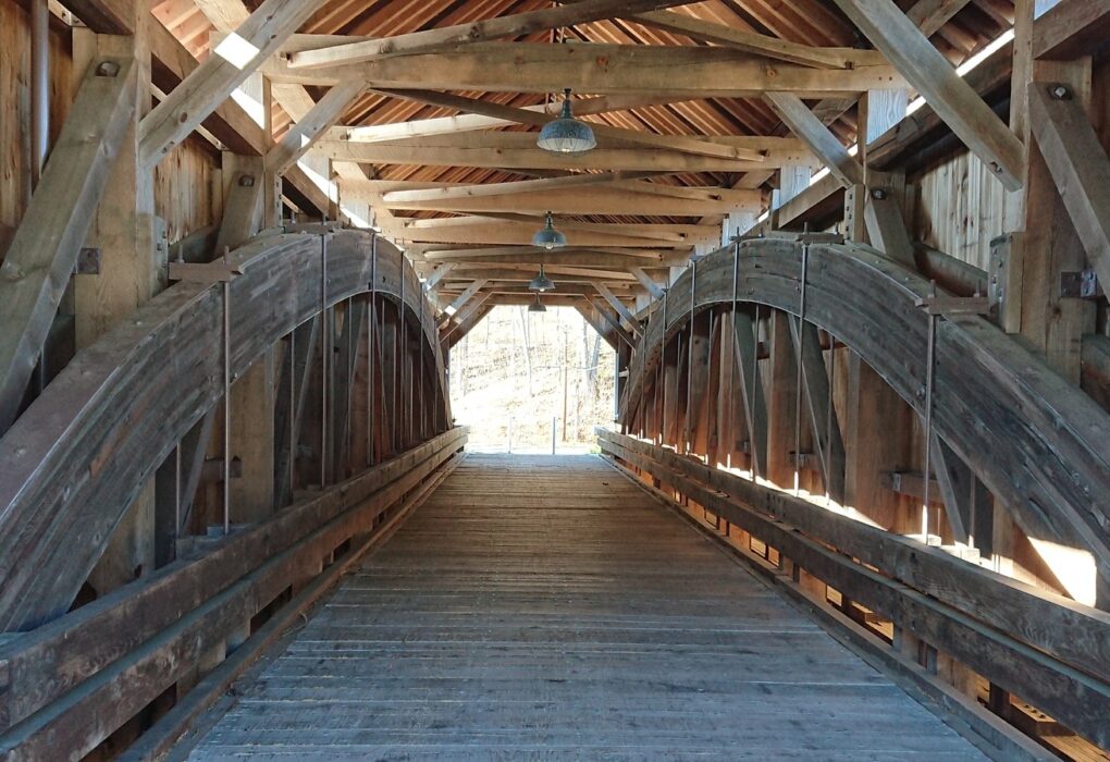 Inside an empty wooden covered bridge: one long timber roof with hanging lamps over a plank deck, a large curved wooden arch truss on each side, and daylight at the far end