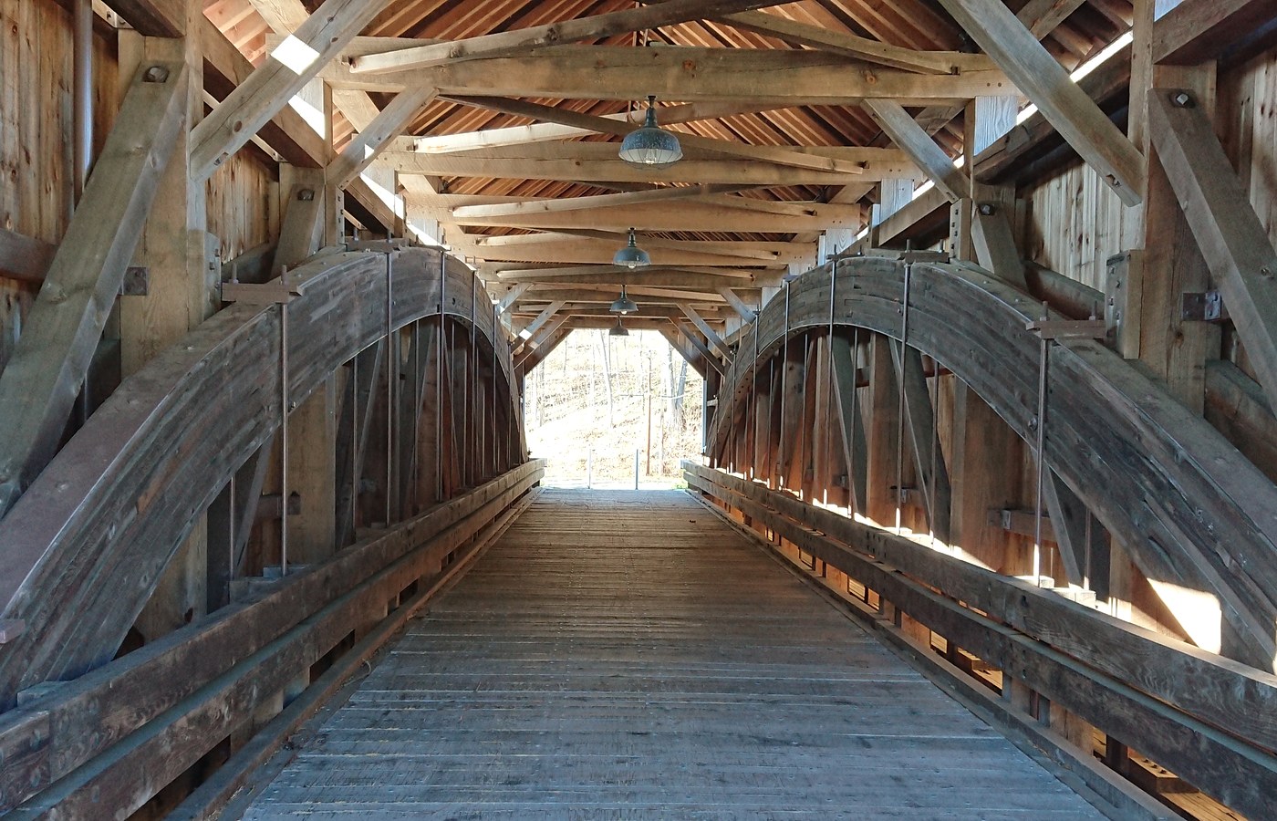 Inside an empty wooden covered bridge: one long timber roof with hanging lamps over a plank deck, a large curved wooden arch truss on each side, and daylight at the far end