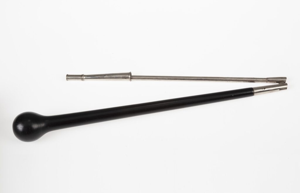A black ebony drum major baton with a round ball head lying beside its separate silver metal shaft on a plain white backdrop