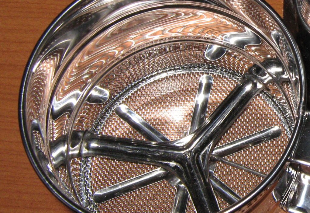 Looking down into a stainless steel flour sifter on a wooden table: fine wire mesh at the bottom, a curved four-armed agitator and a shiny round rim