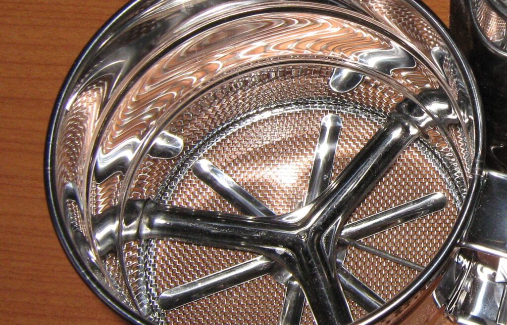 Looking down into a stainless steel flour sifter on a wooden table: fine wire mesh at the bottom, a curved four-armed agitator and a shiny round rim