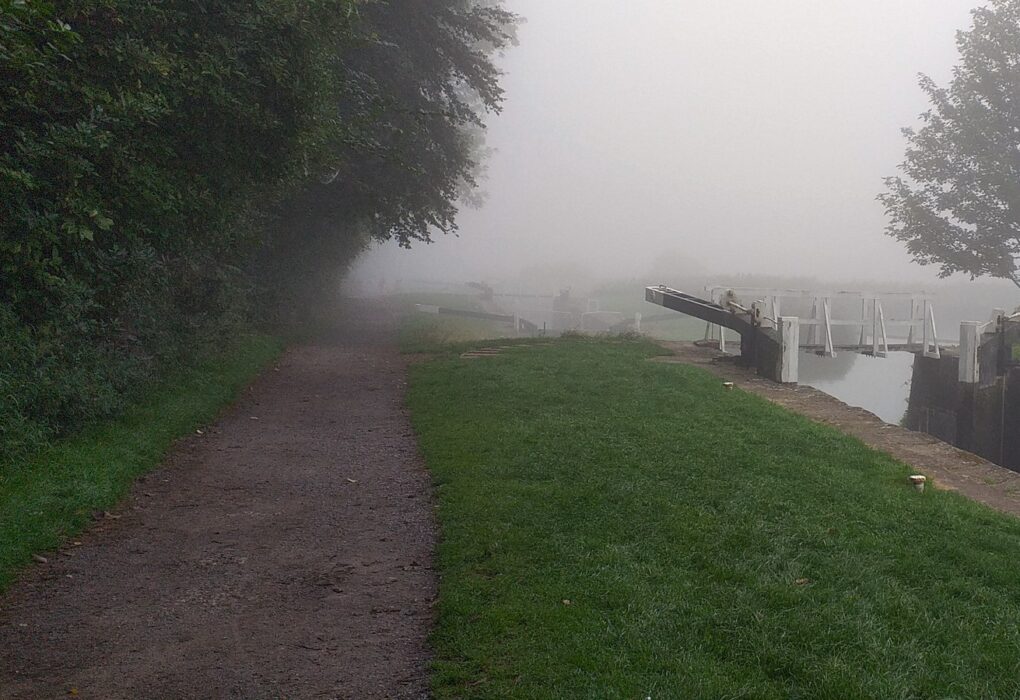 A canal towpath and grassy bank beside a lock gate fading into thick fog, with no people in frame