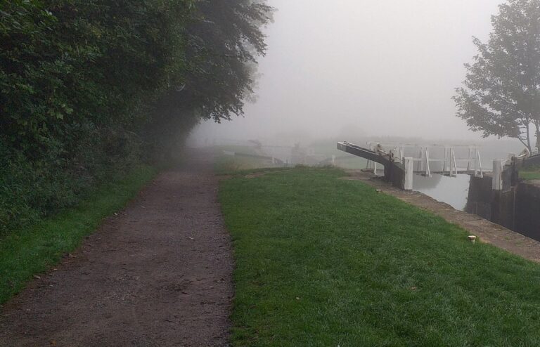 A canal towpath and grassy bank beside a lock gate fading into thick fog, with no people in frame