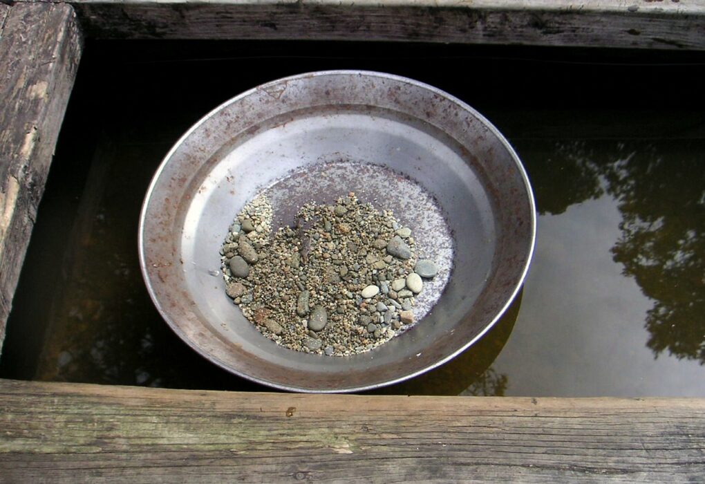 A steel gold pan holding a small pile of wet gravel and pebbles, resting in dark water inside a weathered wooden sluice frame, with no people in frame