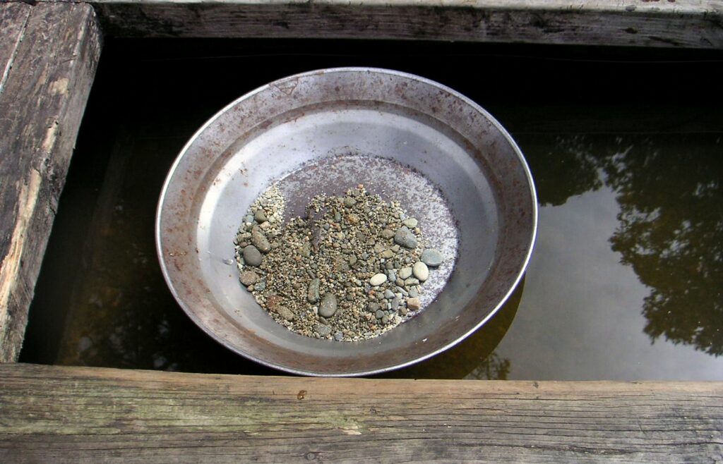 A steel gold pan holding a small pile of wet gravel and pebbles, resting in dark water inside a weathered wooden sluice frame, with no people in frame