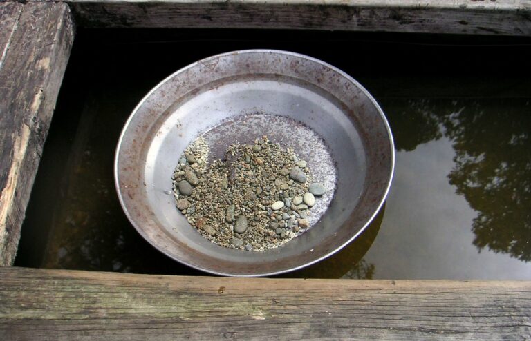 A steel gold pan holding a small pile of wet gravel and pebbles, resting in dark water inside a weathered wooden sluice frame, with no people in frame