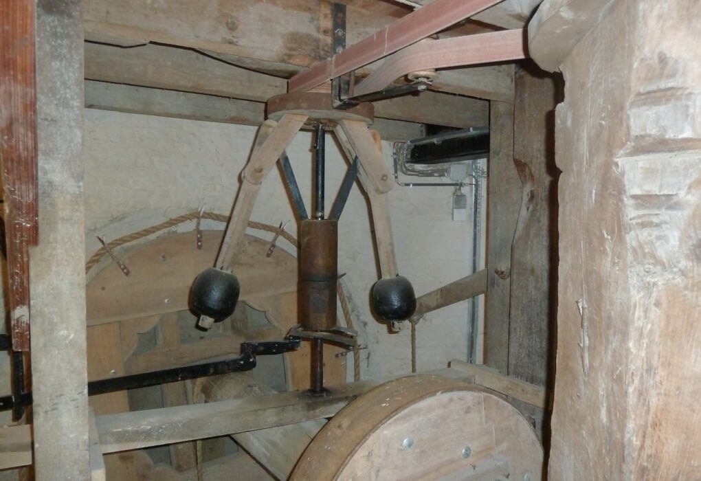 A wooden flyball governor inside an old windmill: two black iron balls on wooden arms around a central spindle, a leather belt overhead and a large wooden wheel below