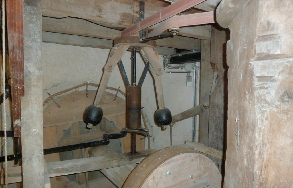 A wooden flyball governor inside an old windmill: two black iron balls on wooden arms around a central spindle, a leather belt overhead and a large wooden wheel below