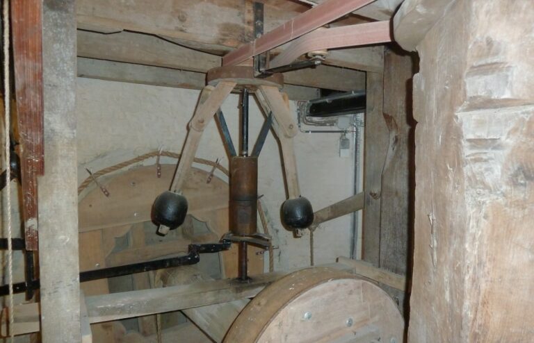 A wooden flyball governor inside an old windmill: two black iron balls on wooden arms around a central spindle, a leather belt overhead and a large wooden wheel below