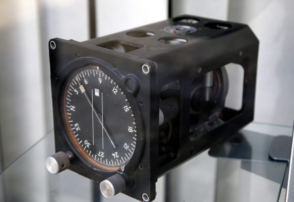 A black aircraft gyrocompass unit in a glass display case, with a round heading dial, a needle and two silver knobs on the front and the gyro mechanism visible through open side panels