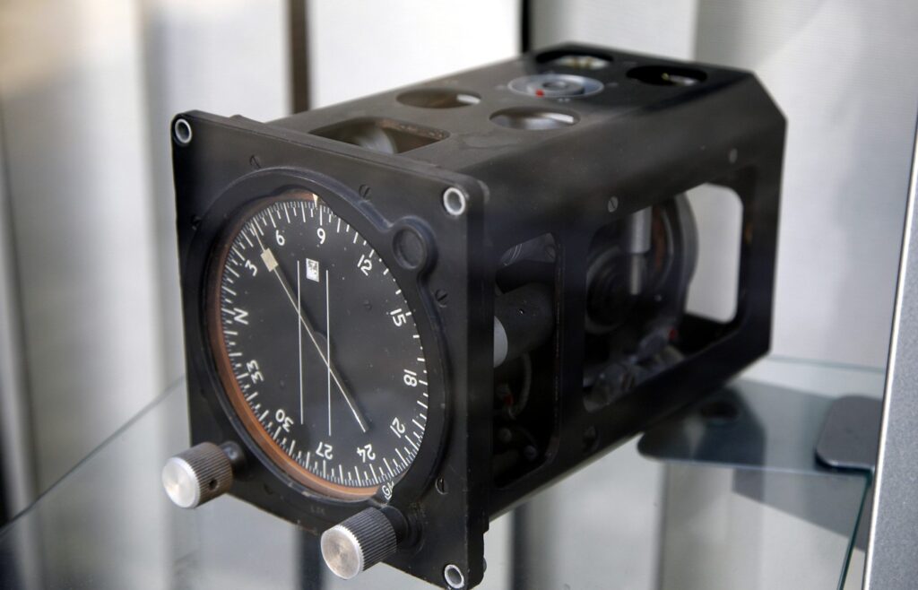 A black aircraft gyrocompass unit in a glass display case, with a round heading dial, a needle and two silver knobs on the front and the gyro mechanism visible through open side panels