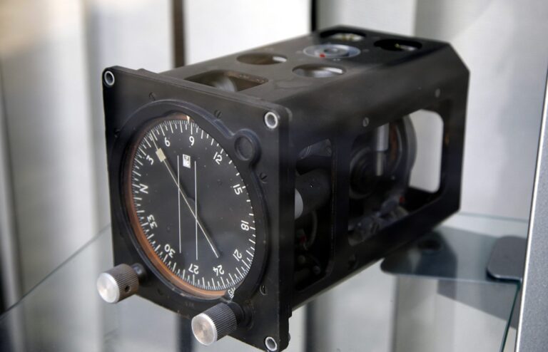 A black aircraft gyrocompass unit in a glass display case, with a round heading dial, a needle and two silver knobs on the front and the gyro mechanism visible through open side panels