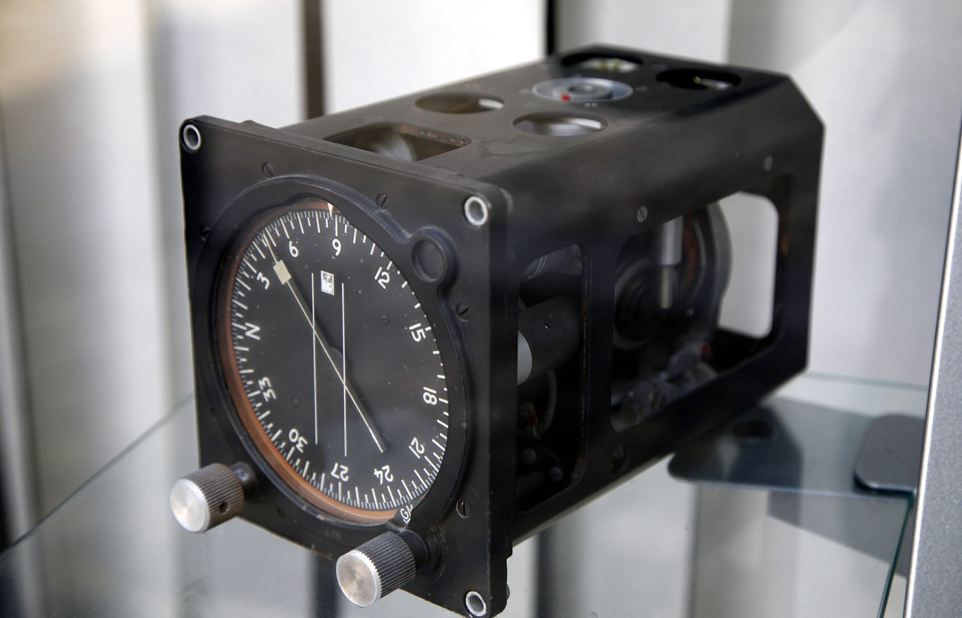 A black aircraft gyrocompass unit in a glass display case, with a round heading dial, a needle and two silver knobs on the front and the gyro mechanism visible through open side panels
