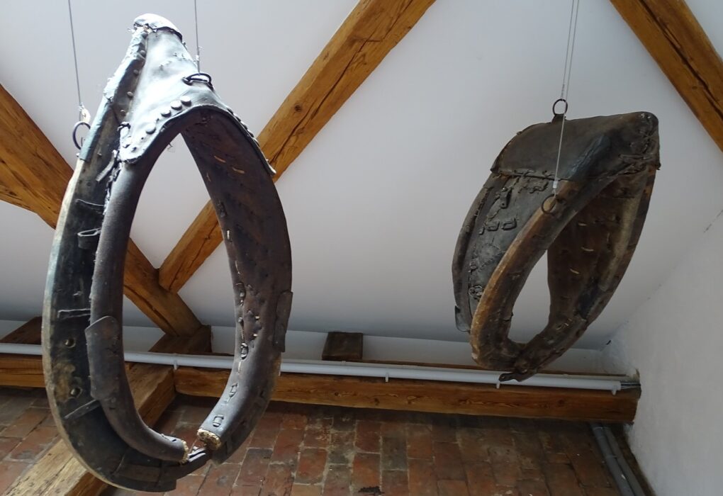 Two old leather-and-wood horse collars hanging from wires under timber roof beams above a brick floor