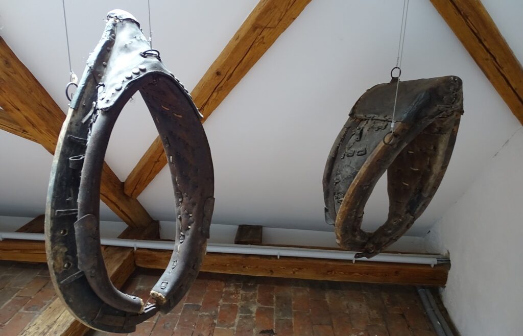 Two old leather-and-wood horse collars hanging from wires under timber roof beams above a brick floor