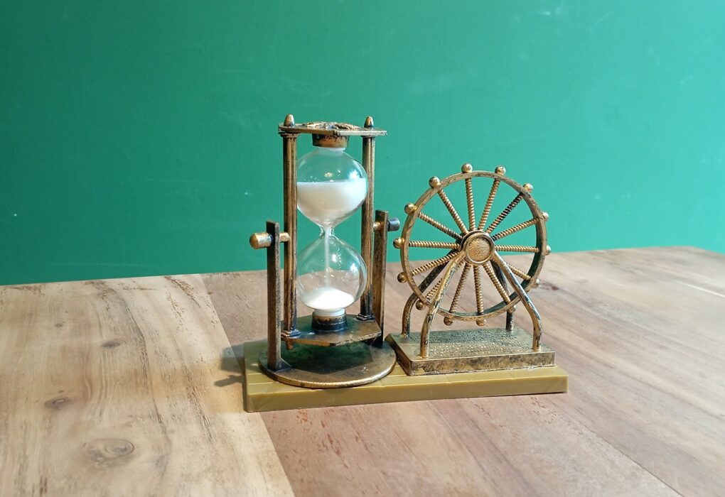 A small brass-colored hourglass with white sand in a pillared frame beside a matching decorative wheel ornament on a wooden table against a plain green wall