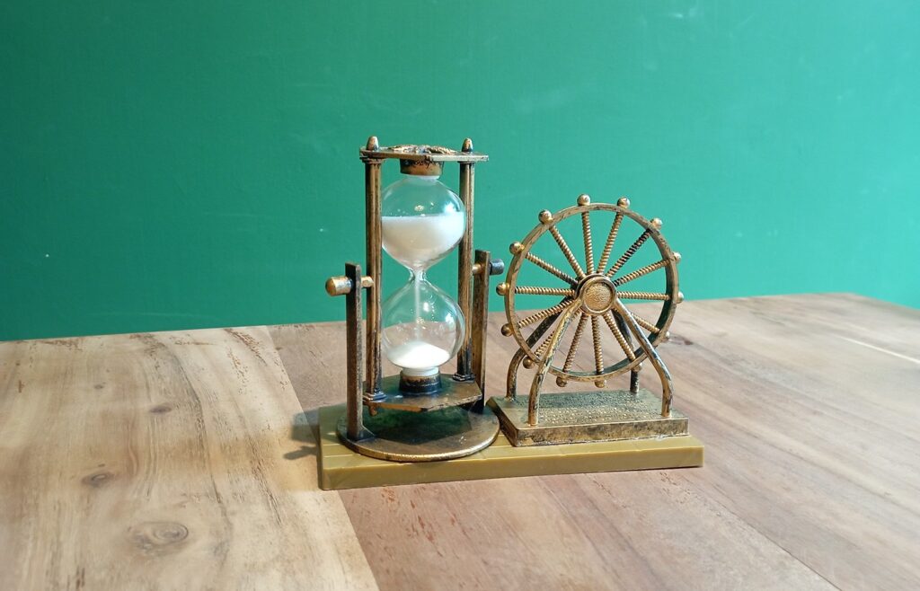 A small brass-colored hourglass with white sand in a pillared frame beside a matching decorative wheel ornament on a wooden table against a plain green wall