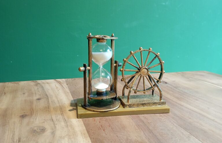 A small brass-colored hourglass with white sand in a pillared frame beside a matching decorative wheel ornament on a wooden table against a plain green wall
