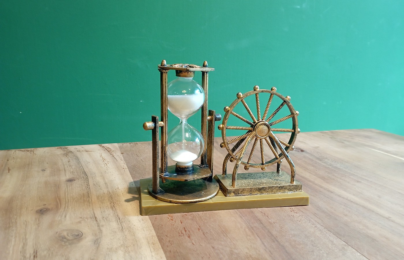 A small brass-colored hourglass with white sand in a pillared frame beside a matching decorative wheel ornament on a wooden table against a plain green wall