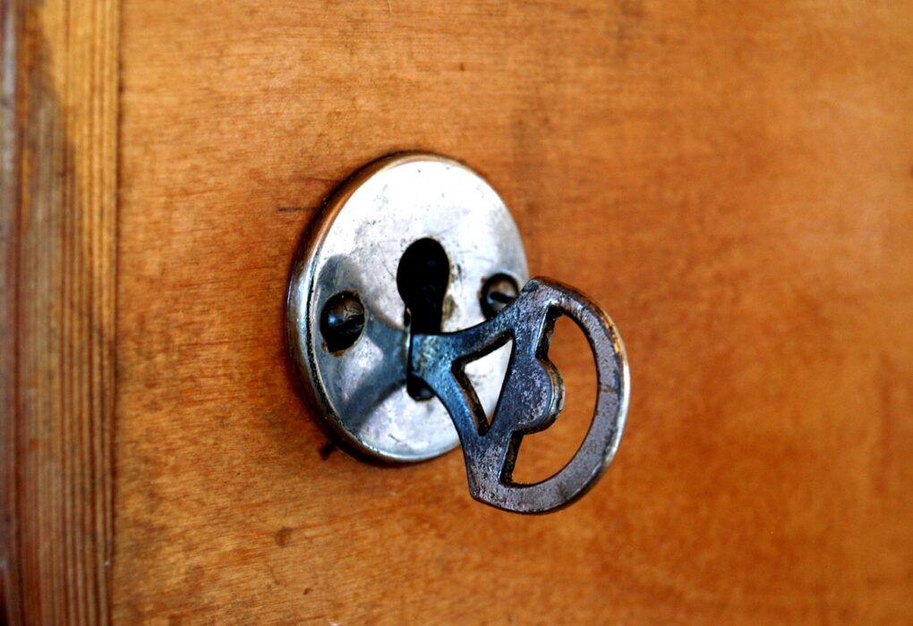 An old iron key left in a round, worn metal escutcheon on a warm wooden door, with a vertical door frame on the left and no people in frame