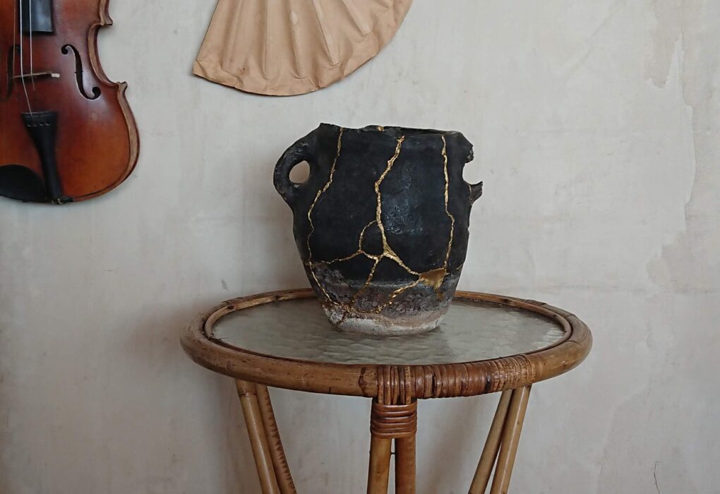 A dark old cooking pot with its cracks rejoined in bright gold lines, on a rattan side table against a pale plaster wall