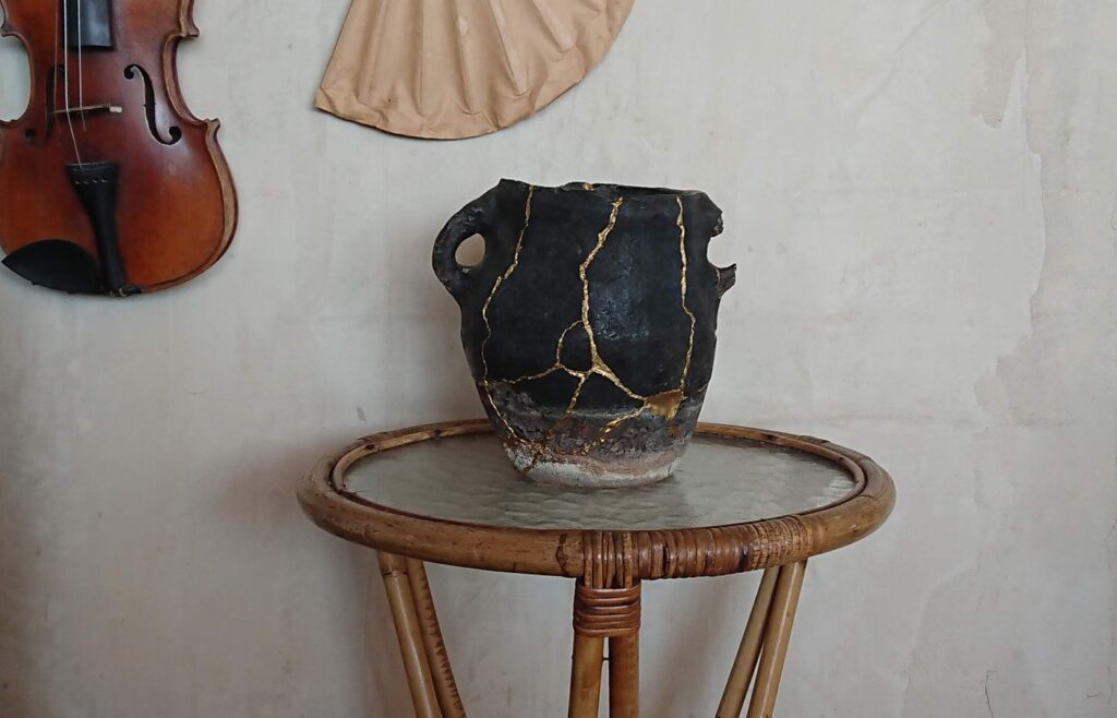 A dark old cooking pot with its cracks rejoined in bright gold lines, on a rattan side table against a pale plaster wall
