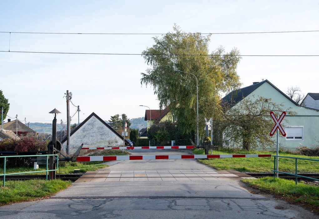 A lowered red-and-white level-crossing barrier across a quiet village road, with houses, a tall tree and a railway crossing sign